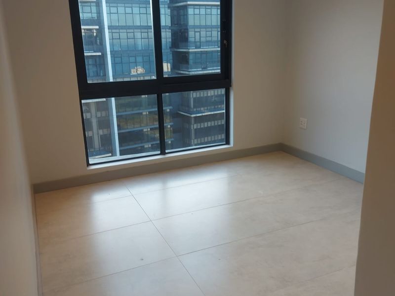 A bright, empty bedroom featuring neutral tiled flooring, modern recessed ceiling lighting, and a large window with a city view.
