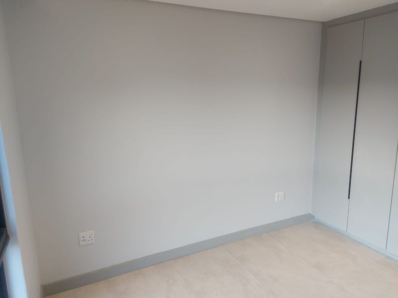 A clean, modern bedroom featuring neutral grey walls, tiled flooring, and built-in floor-to-ceiling cupboards with sleek vertical handles.