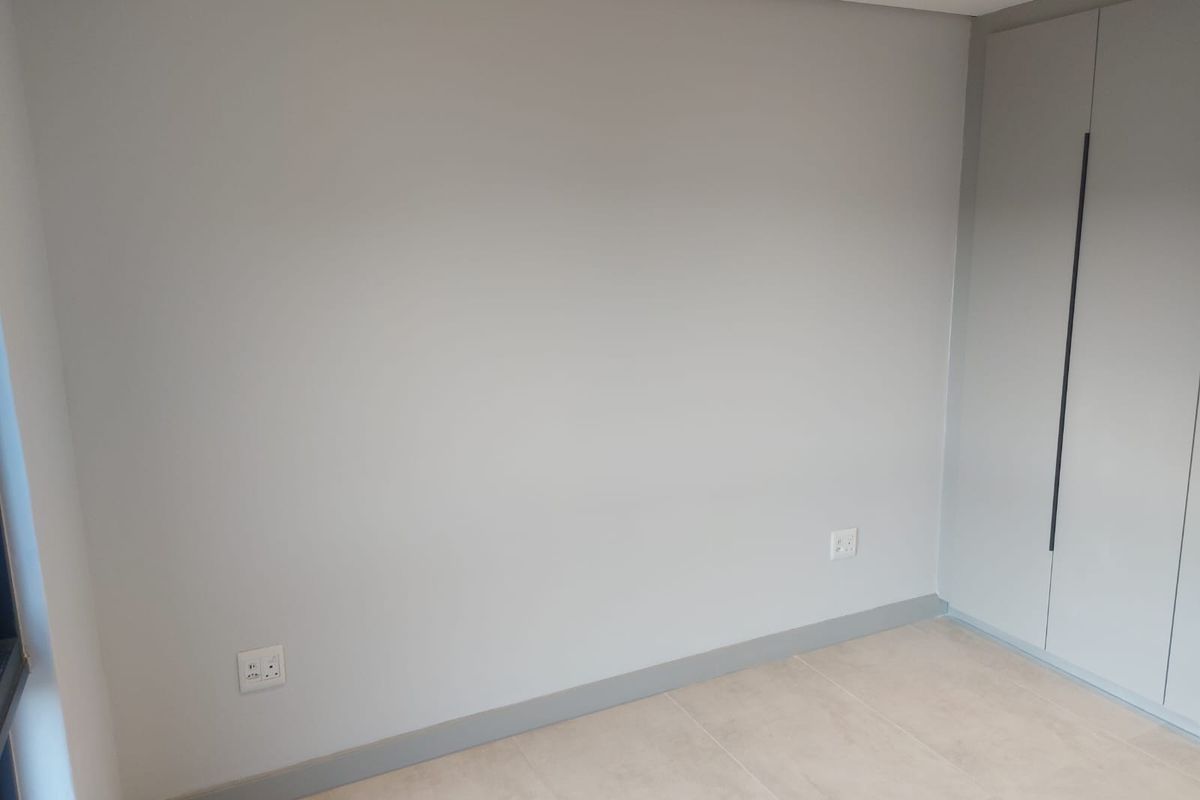 A clean, modern bedroom featuring neutral grey walls, tiled flooring, and built-in floor-to-ceiling cupboards with sleek vertical handles.