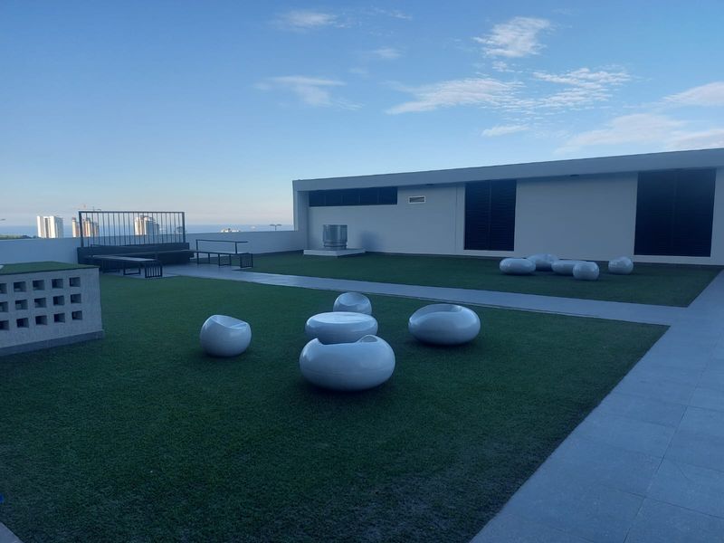 A spacious rooftop terrace featuring artificial turf, paved walkways, and metal safety railings, offering an open-air communal space with distant city and sea views.