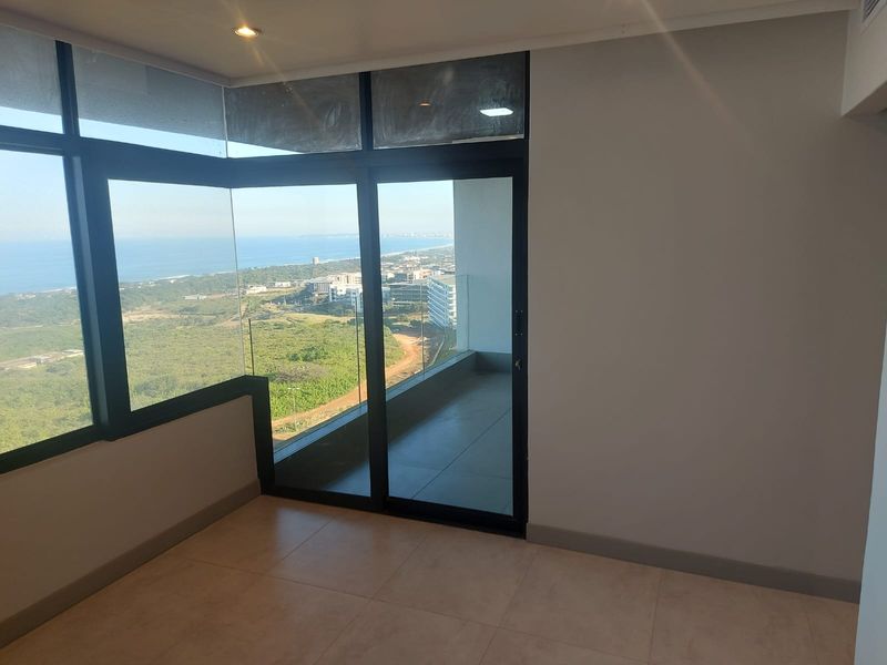 A bright, empty living area featuring large floor-to-ceiling windows and a sliding glass door leading to a private balcony with expansive coastal views.