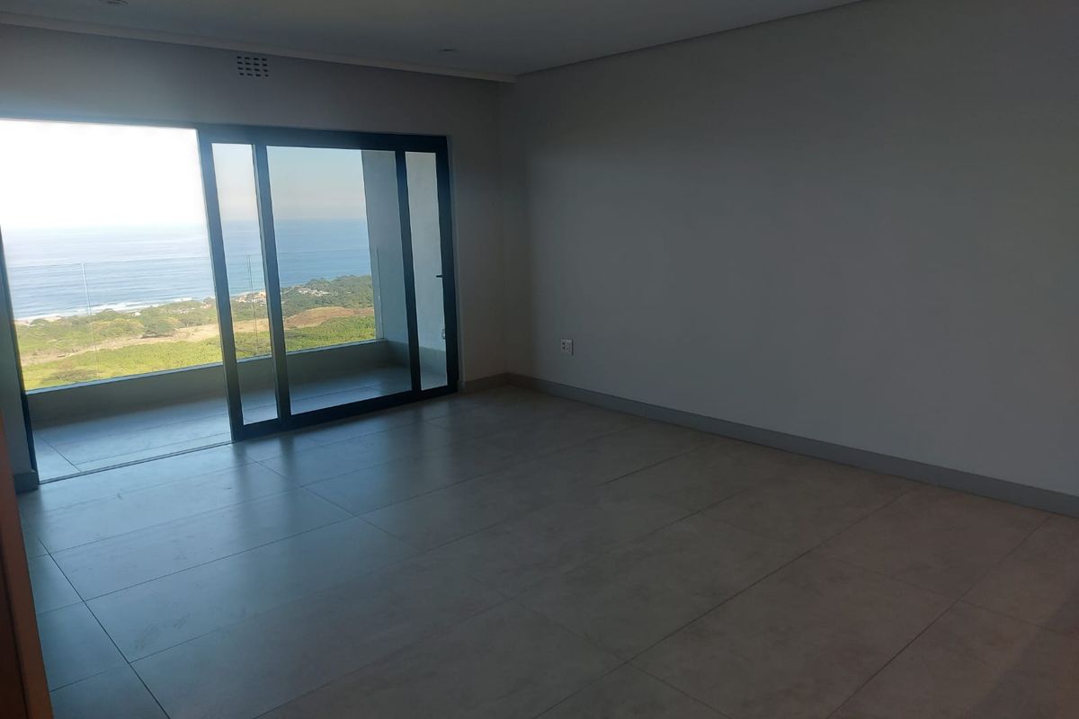 Spacious, empty living area featuring large sliding glass doors that open onto a balcony with expansive ocean and coastal views.