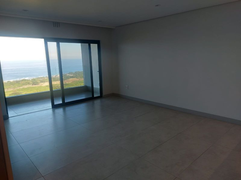 Spacious, empty living area featuring large sliding glass doors that open onto a balcony with expansive ocean and coastal views.