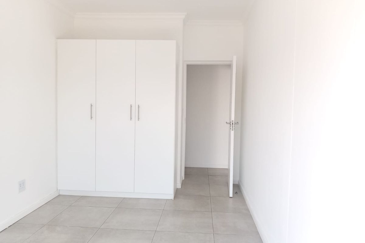 A bright, minimalist bedroom featuring neutral floor tiles and a built-in white wardrobe with modern handles.
