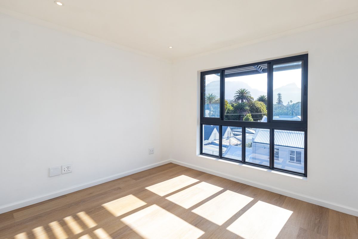 A bright, empty bedroom featuring modern light wood-look flooring, recessed ceiling lighting, and a large window with dark frames offering a scenic neighborhood view.