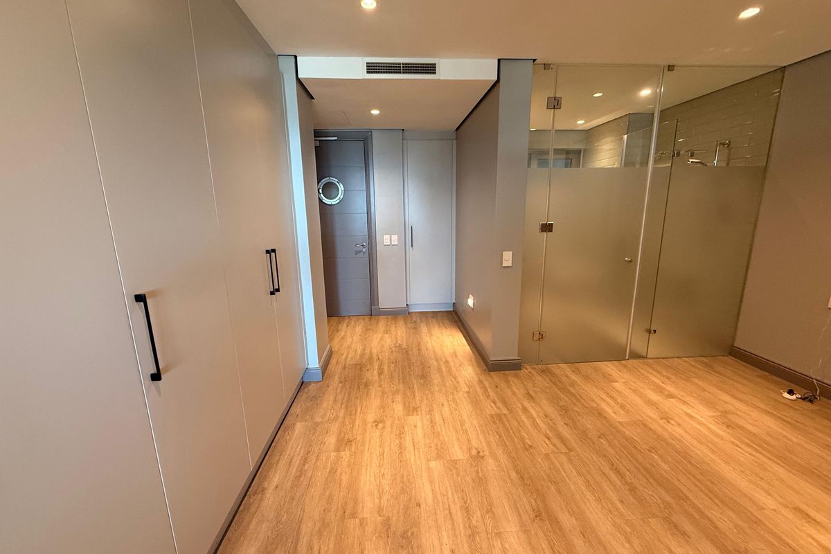 A modern bedroom interior featuring built-in cabinetry, wood-look flooring, recessed ceiling lighting, and an en-suite bathroom with a glass partition.