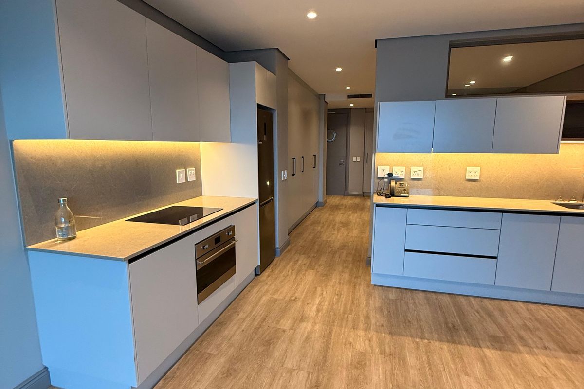 Modern kitchen featuring sleek cabinetry, integrated oven and hob, stone countertops, and warm under-cabinet lighting, set against light wood-look flooring.