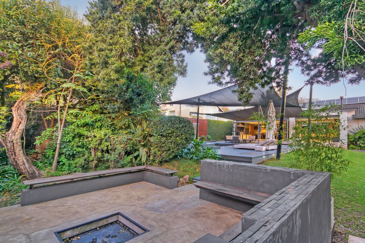 A landscaped rear garden featuring a sunken fire pit area with built-in concrete seating, mature trees, and a raised deck with shade sails.