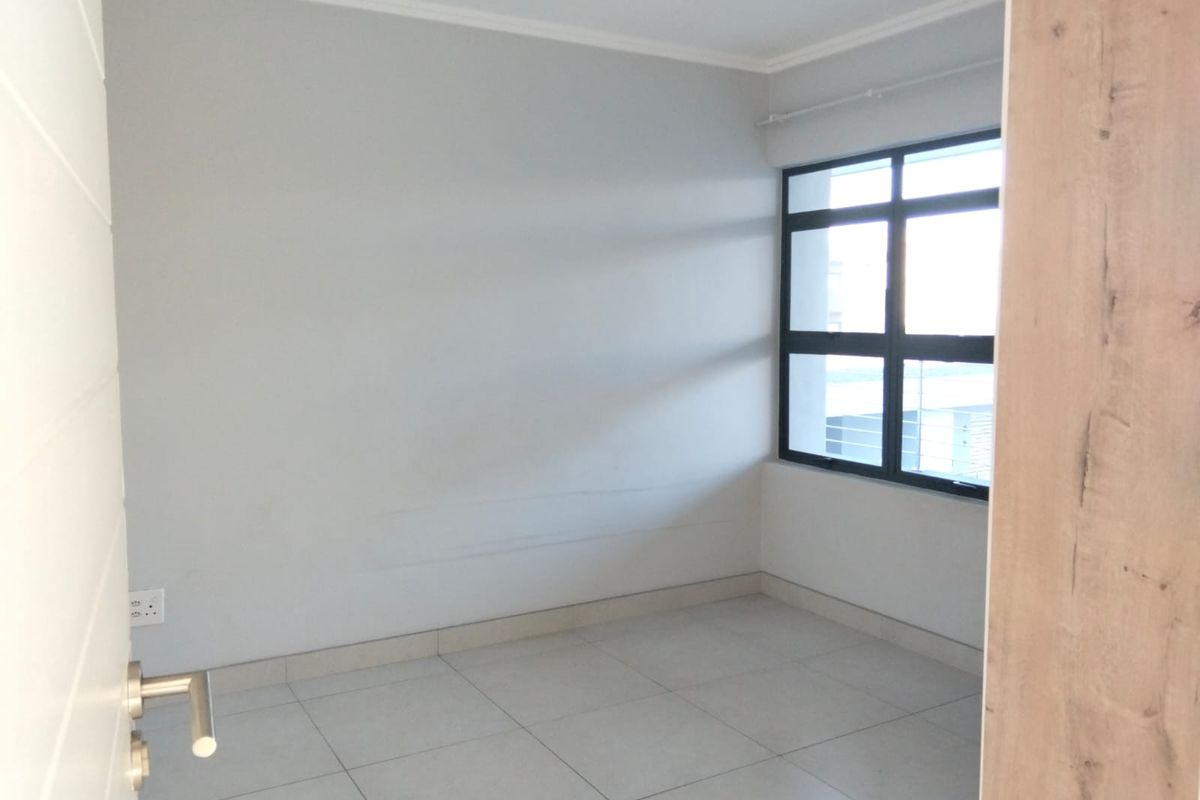 A bright, empty bedroom featuring neutral tiled flooring, clean walls, and a large aluminium-framed window providing natural light.