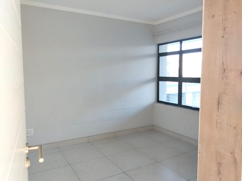 A bright, empty bedroom featuring neutral tiled flooring, clean walls, and a large aluminium-framed window providing natural light.