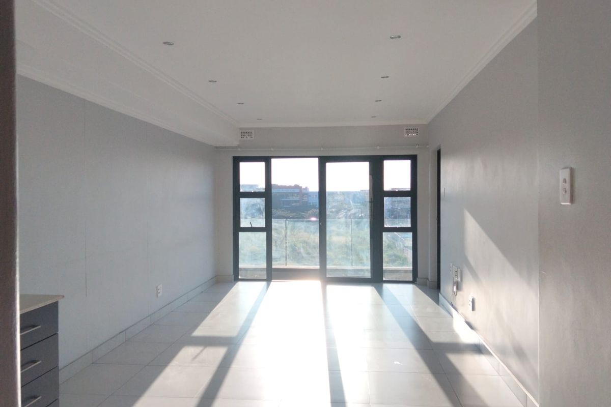 A bright, empty living area featuring neutral tiled flooring, recessed lighting, and large sliding glass doors leading to a balcony with a view.
