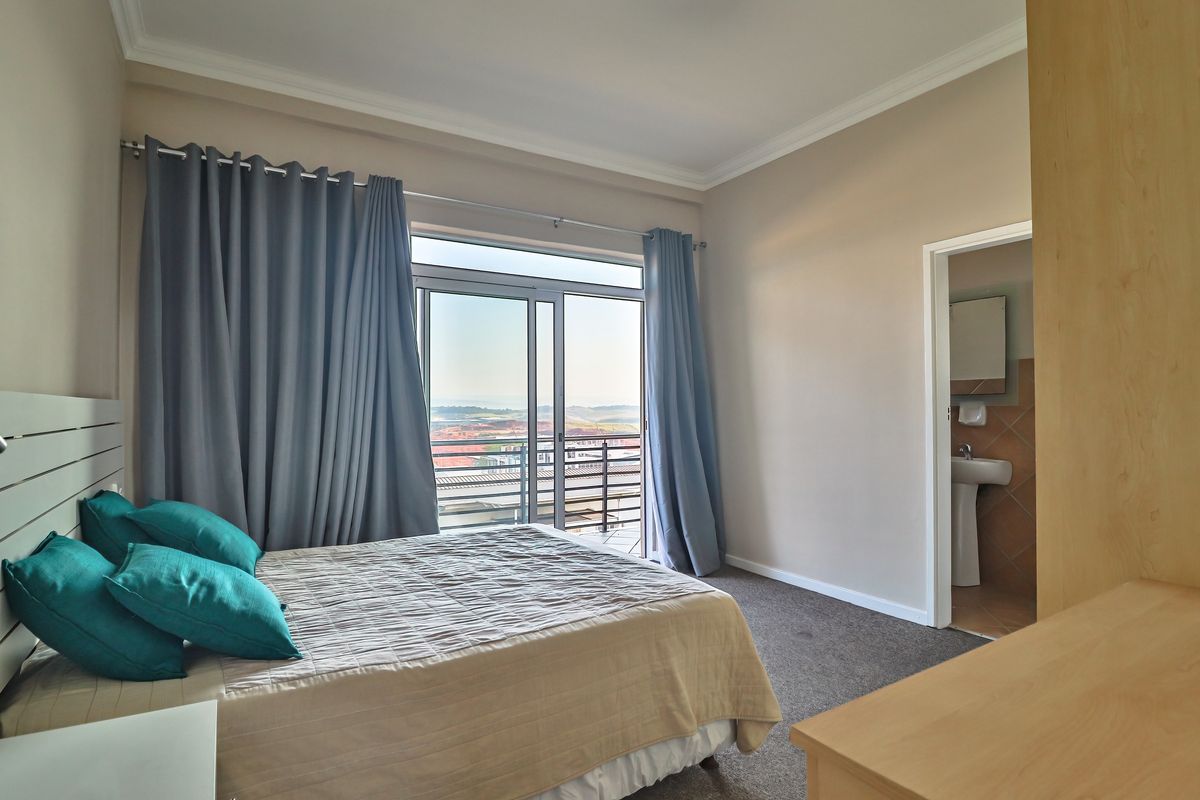 A well-lit bedroom featuring neutral walls, carpeted flooring, a sliding door leading to a balcony, and an attached en-suite bathroom with a pedestal basin.