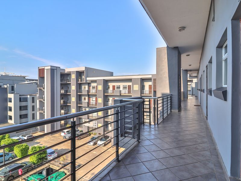 A spacious covered balcony featuring tiled flooring and a metal balustrade, offering an elevated view of the apartment complex and parking area.