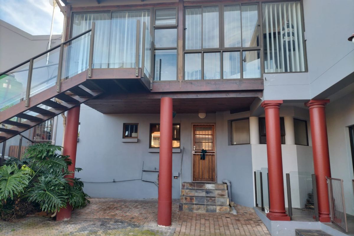 Rear exterior view featuring a raised timber balcony with glass balustrades, supported by red columns, leading to a paved patio area.