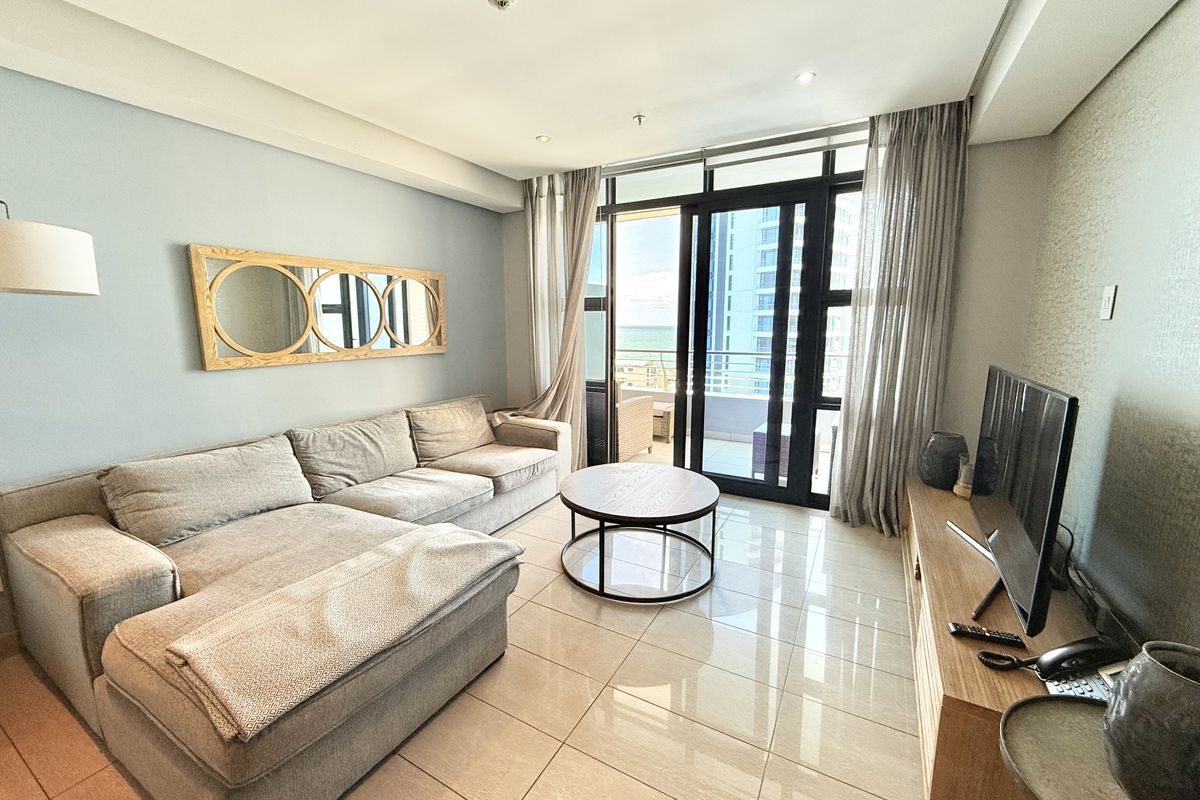 A bright and modern living room featuring large sliding glass doors that open onto a balcony with coastal views and polished floor tiles.