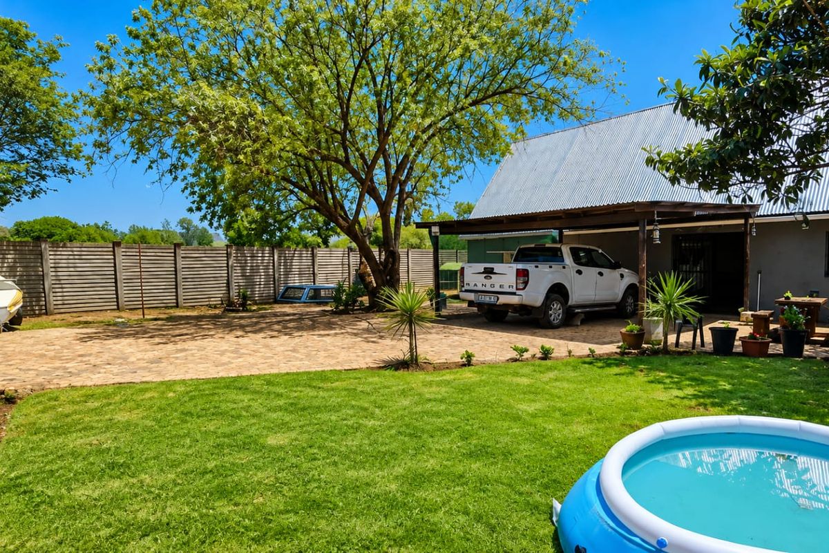 Rear exterior view of the small holding featuring a paved driveway, covered patio area, mature trees, and a well-maintained lawn.