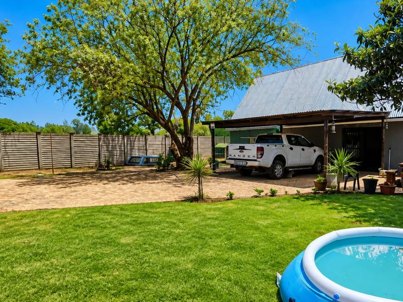 Rear exterior view of the small holding featuring a paved driveway, covered patio area, mature trees, and a well-maintained lawn.