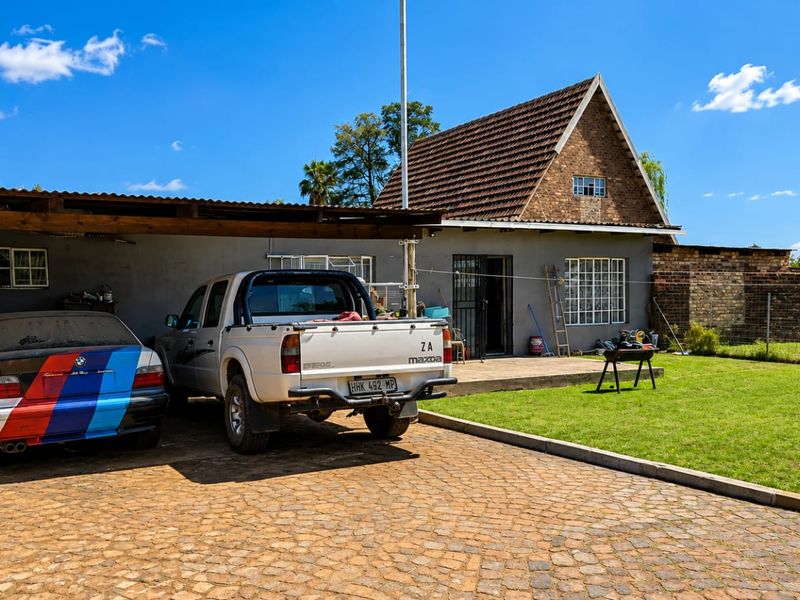 Rear view of the property featuring a carport, paved driveway, lawn area, and a brick-built structure with a tiled roof.