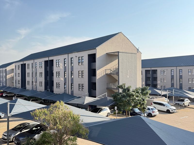 Exterior view of a modern apartment complex featuring multi-level residential blocks, paved parking areas with shade cloth carports, and landscaped grounds.