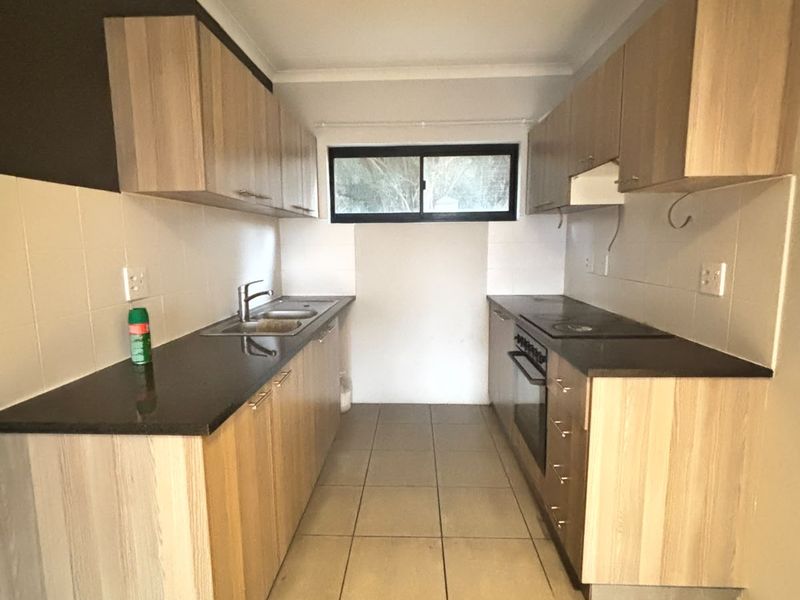A galley-style kitchen featuring light wood-finish cabinetry, dark countertops, built-in oven and hob, and neutral tiled flooring.