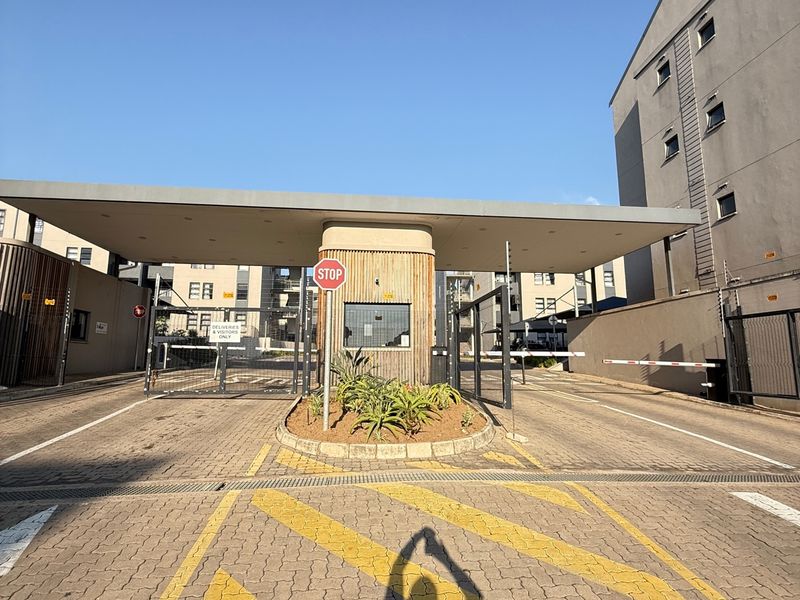 The main entrance to the apartment complex featuring a secure guard house, boom gate access, and paved driveway.