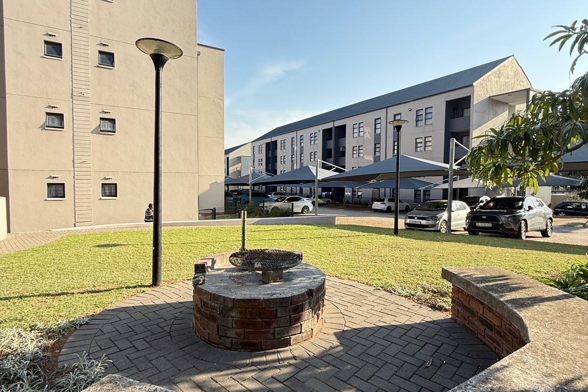Communal outdoor area featuring a brick braai, paved seating space with a curved concrete bench, and well-maintained lawn within an apartment complex.