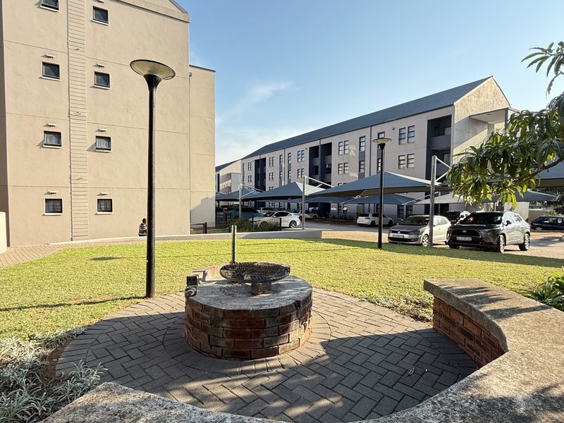 Communal outdoor area featuring a brick braai, paved seating space with a curved concrete bench, and well-maintained lawn within an apartment complex.