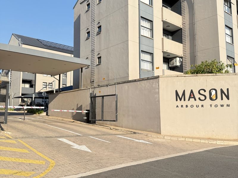 The entrance to Mason Arbour Town apartment complex, featuring a secure boom gate, paved driveway, and modern multi-story residential building facade.