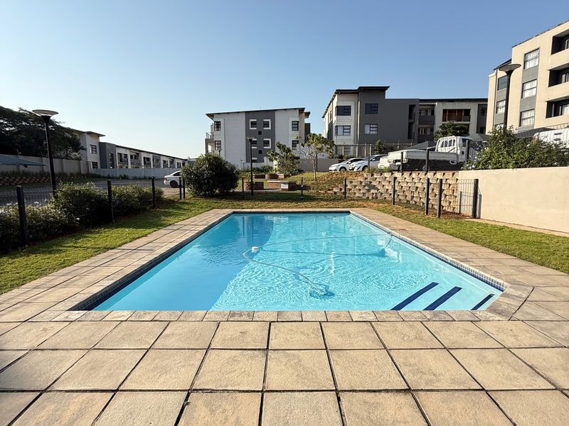 A well-maintained outdoor swimming pool area featuring a paved deck, perimeter fencing, and landscaped surroundings within an apartment complex.
