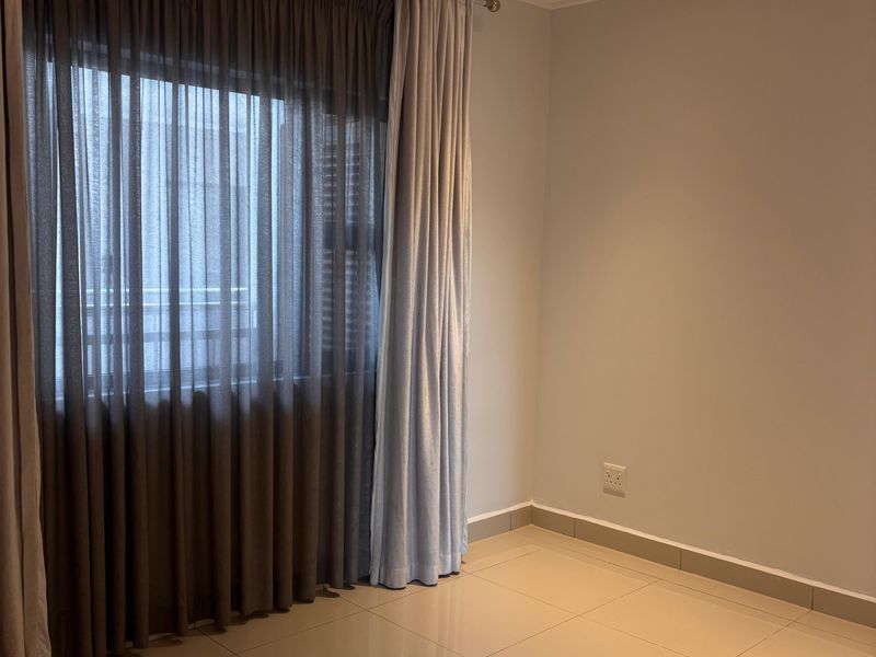 A bright, empty bedroom featuring neutral tiled flooring, recessed ceiling lighting, and floor-to-ceiling curtains.