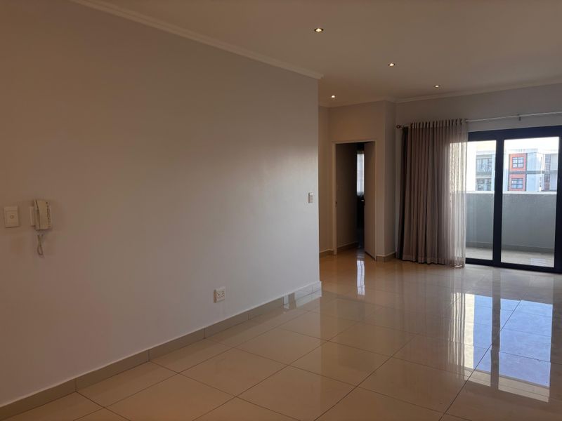 A bright, open-plan living area featuring polished floor tiles, recessed downlighting, and a sliding glass door leading to a private balcony.
