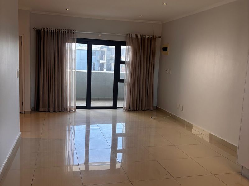 A spacious, empty living area featuring polished floor tiles, recessed downlighting, and sliding glass doors leading to a private balcony.