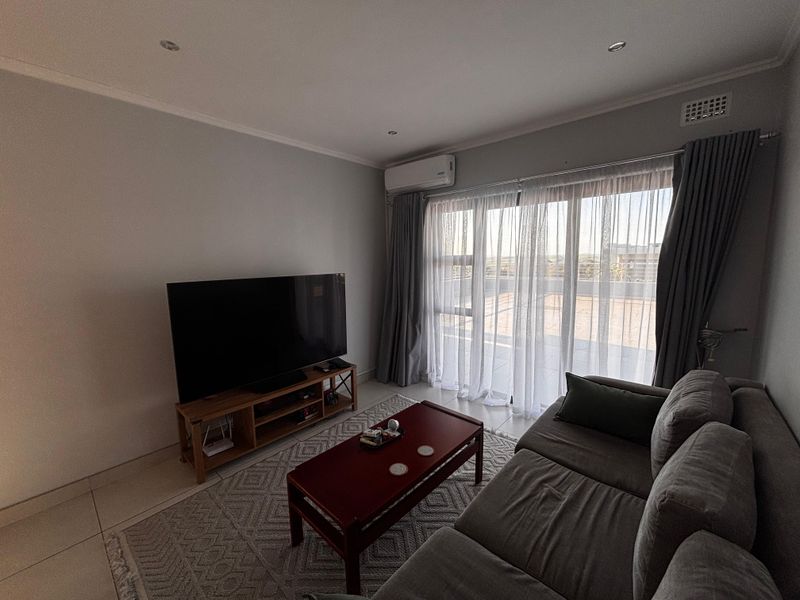 A comfortable living room featuring tiled flooring, recessed downlighting, a wall-mounted air conditioning unit, and large sliding glass doors leading to a balcony.