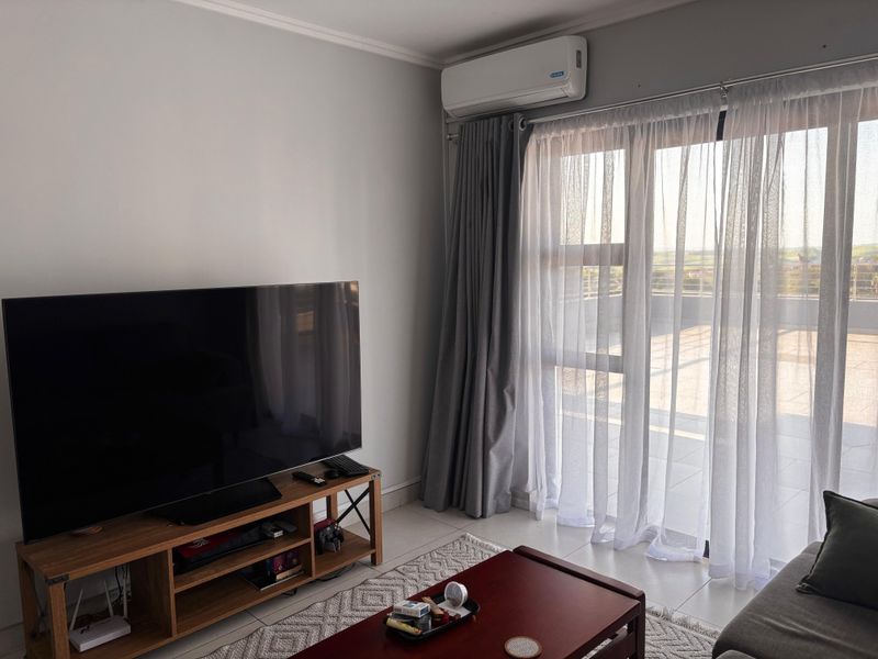 A bright living room featuring tiled flooring, a wall-mounted air conditioning unit, and large sliding glass doors providing access to an outdoor balcony.