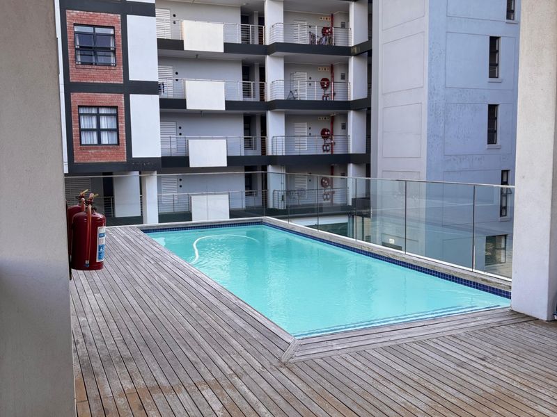 A communal swimming pool area featuring timber decking and a glass balustrade, set within an apartment complex courtyard.