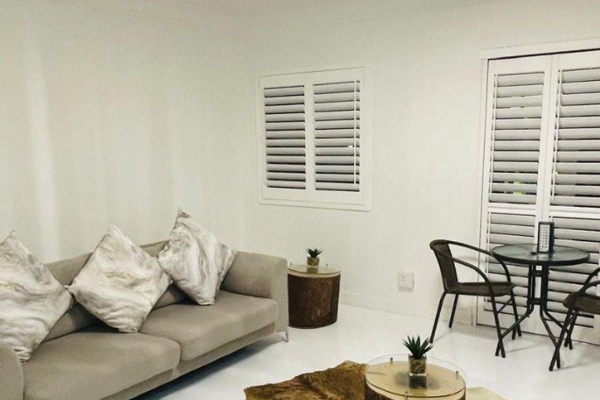 A bright living room featuring white flooring, plantation shutters on the windows and door, and a modern flush-mount ceiling light fixture.