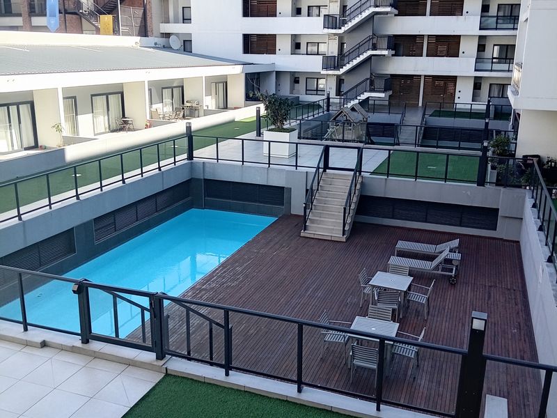 A well-maintained apartment complex courtyard featuring a swimming pool, timber decking, artificial grass areas, and external staircases.