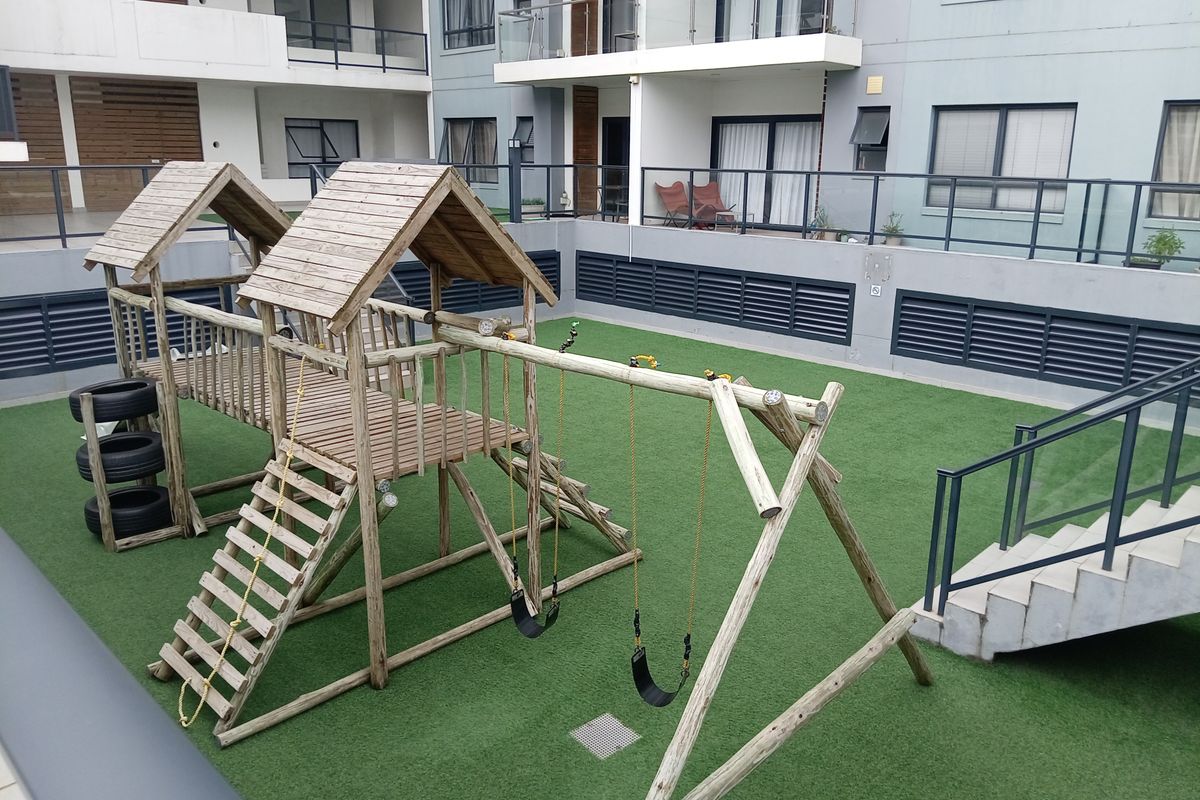Communal outdoor play area featuring a wooden jungle gym set with swings, situated on an artificial grass surface within the apartment complex.