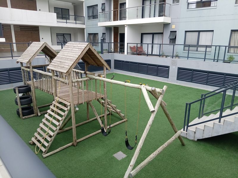 Communal outdoor play area featuring a wooden jungle gym set with swings, situated on an artificial grass surface within the apartment complex.