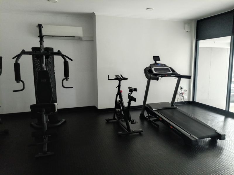 A well-maintained gym space featuring rubber flooring, a wall-mounted air conditioning unit, and large windows providing natural light.