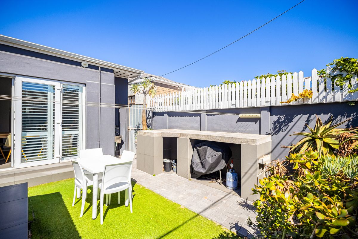 A private rear courtyard featuring a built-in concrete braai area, artificial lawn, and secure perimeter fencing with white picket detail.