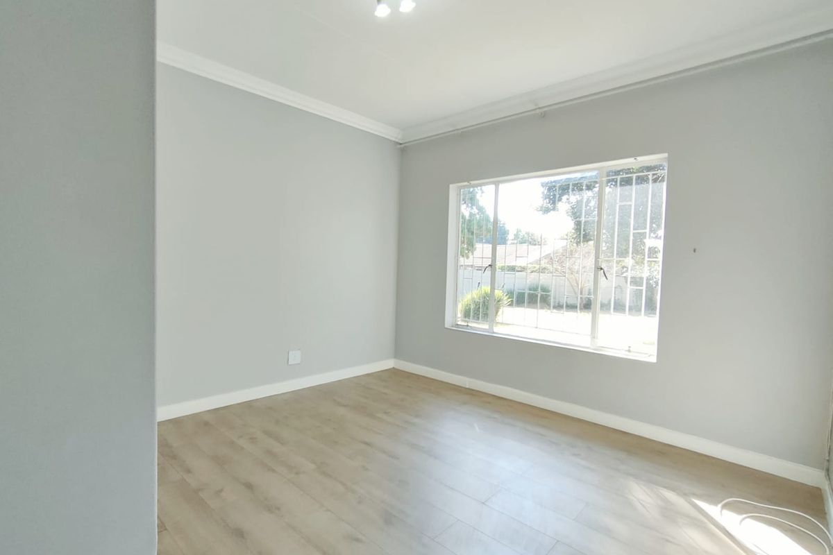 A bright, empty bedroom featuring light-toned laminate flooring, neutral grey walls, and a large window fitted with security bars.