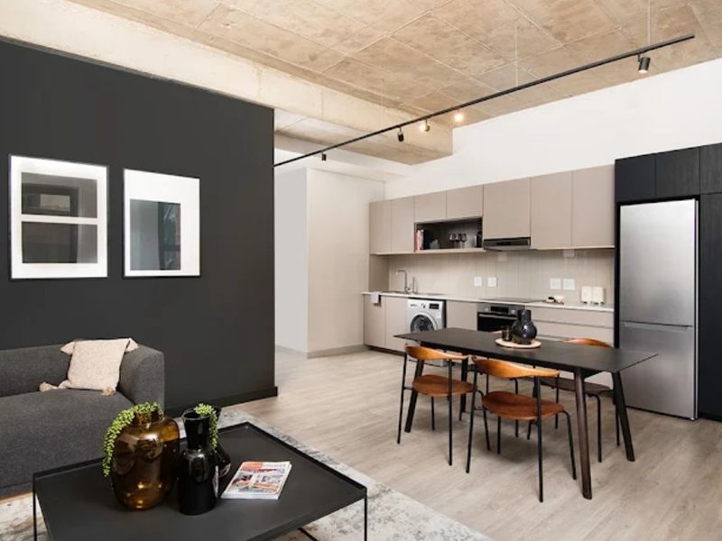 Modern open-plan living area featuring an exposed concrete ceiling, track lighting, and a sleek, contemporary kitchen with integrated appliances.