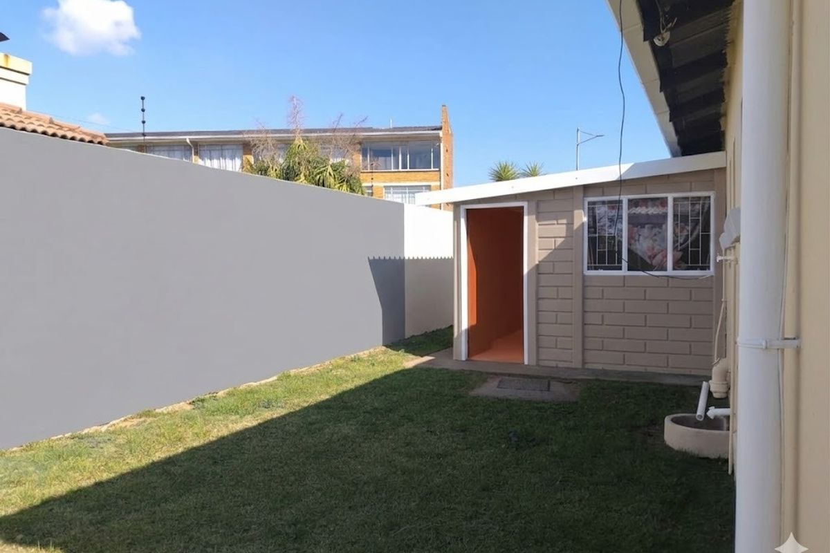 A view of the rear exterior area featuring a small outbuilding, a grass lawn, and a high perimeter boundary wall.