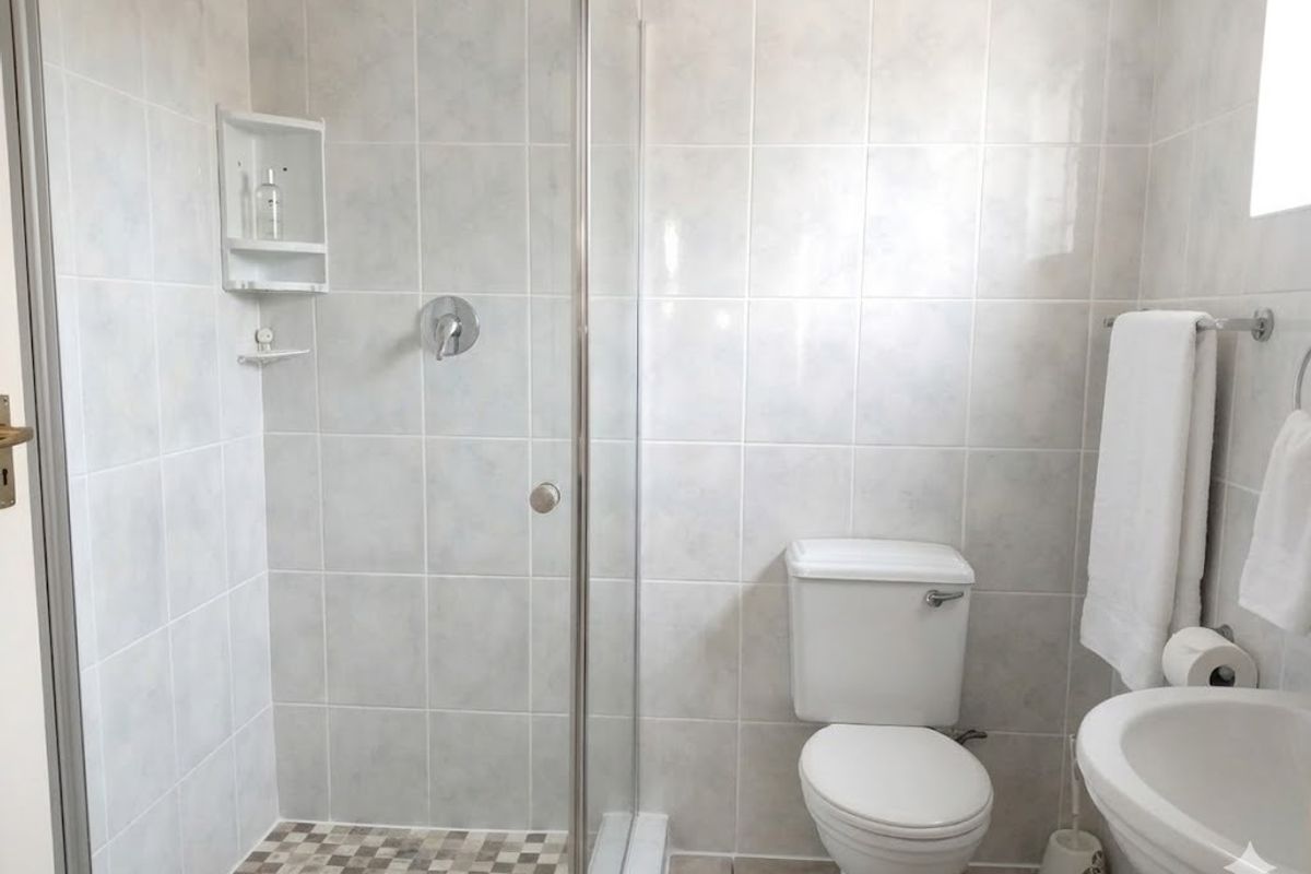 A functional bathroom featuring a glass-enclosed shower, a standard toilet, and a pedestal basin with neutral wall tiling.