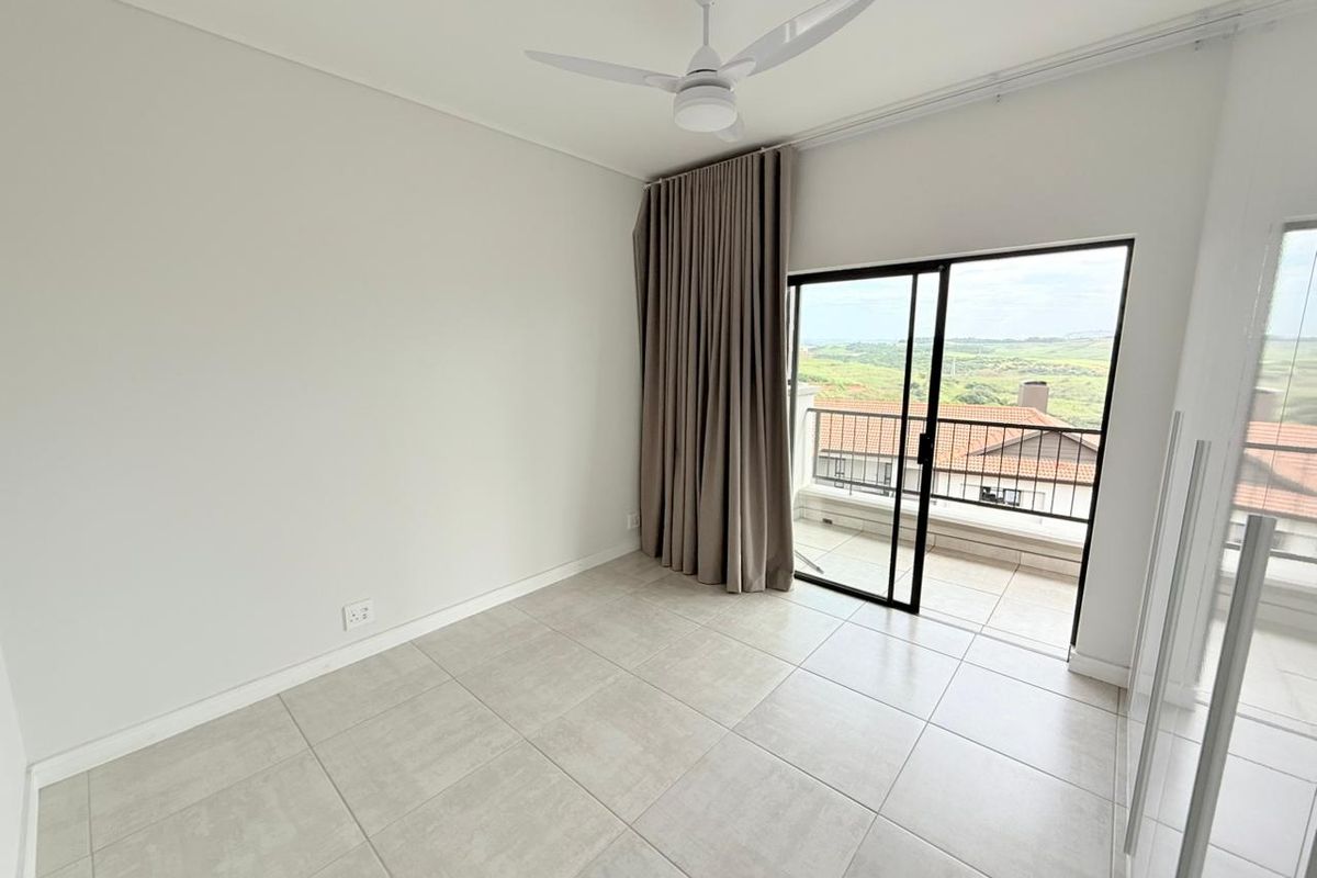 A bright, empty bedroom featuring neutral floor tiles, a ceiling fan, and sliding glass doors opening onto a private balcony with scenic views.