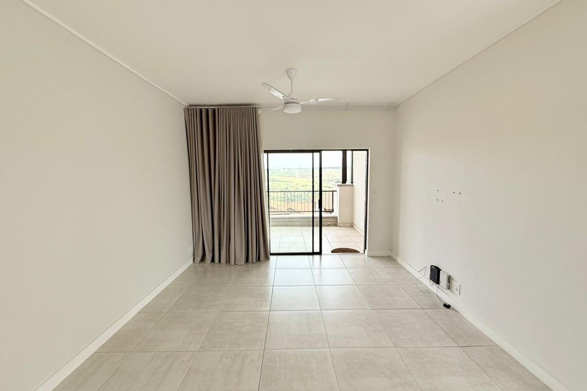 A bright, empty living room featuring neutral floor tiles, a ceiling fan, and sliding glass doors leading to a balcony with scenic views.