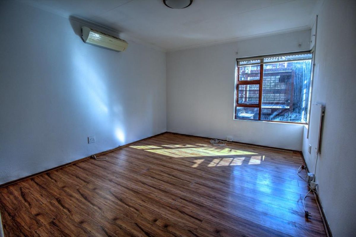 A bright, empty bedroom featuring wood-look laminate flooring, a wall-mounted air conditioning unit, and a window with security bars.