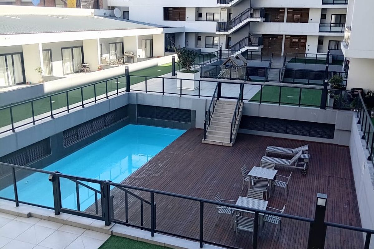 A well-maintained apartment complex courtyard featuring a swimming pool, wooden decking, artificial grass areas, and modern metal balustrades.