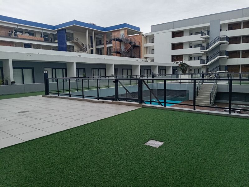 A modern apartment complex courtyard featuring a tiled patio area, artificial turf, glass balustrades, and a sunken swimming pool.